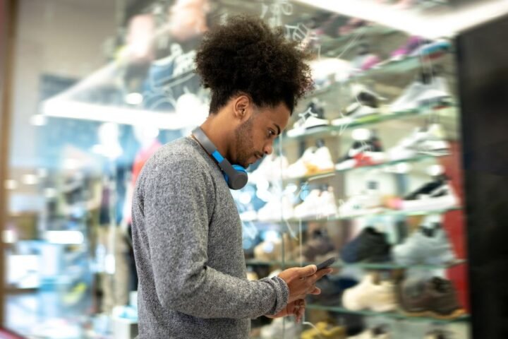 Man looks at phone while standing in front of a store window display of sports shoes