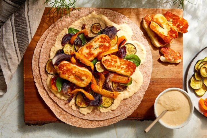 Halloumi and roasted red onion, courgette and bell peppers arranged on a wrap, next to a plate of roasted vegetables and a bowl of hummus
