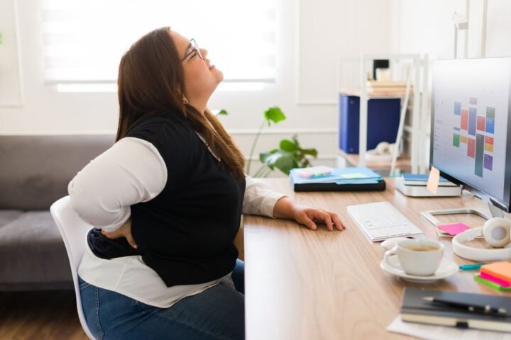 Woman sitting at desk holds her lower back and winces in pain